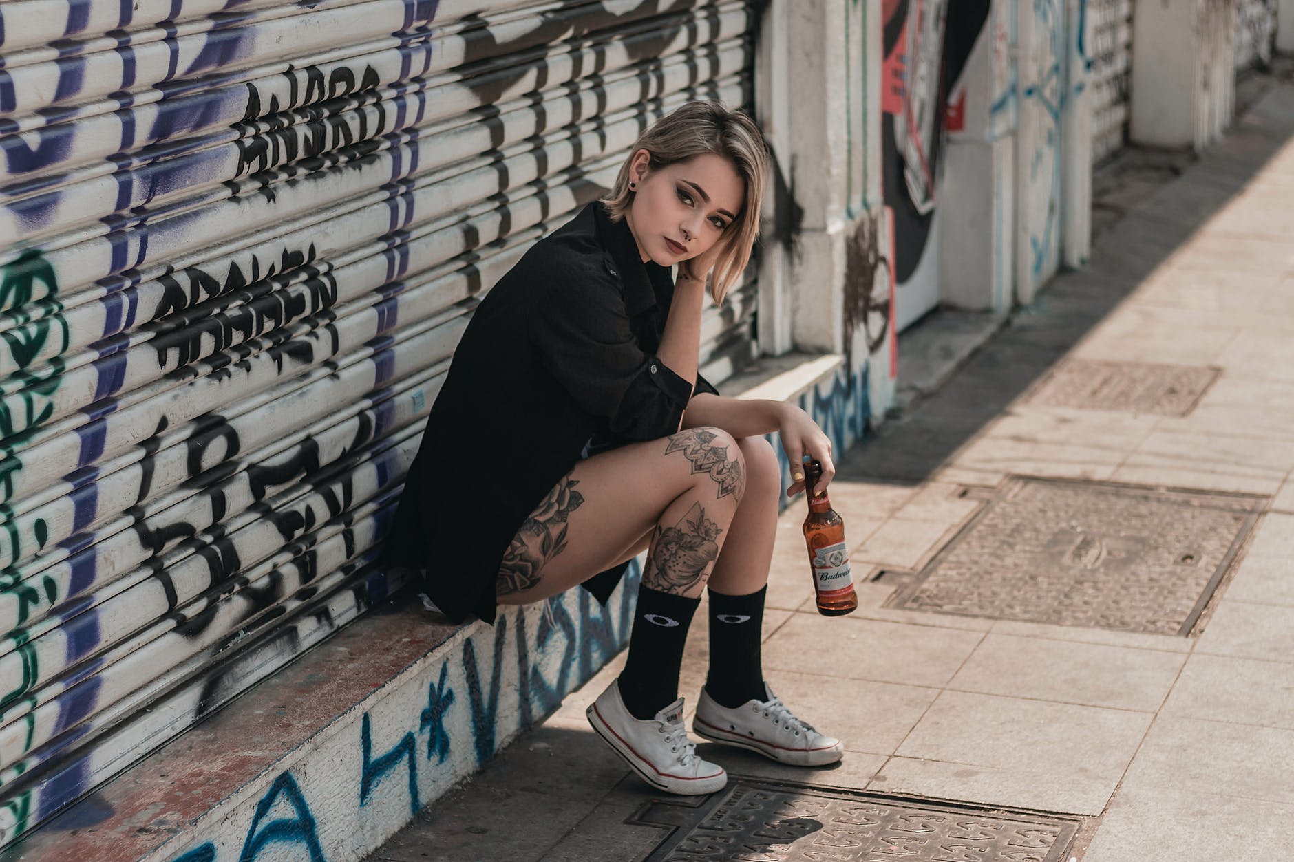 photo of tattooed woman sitting in front of rolling steel door holding beer bottle